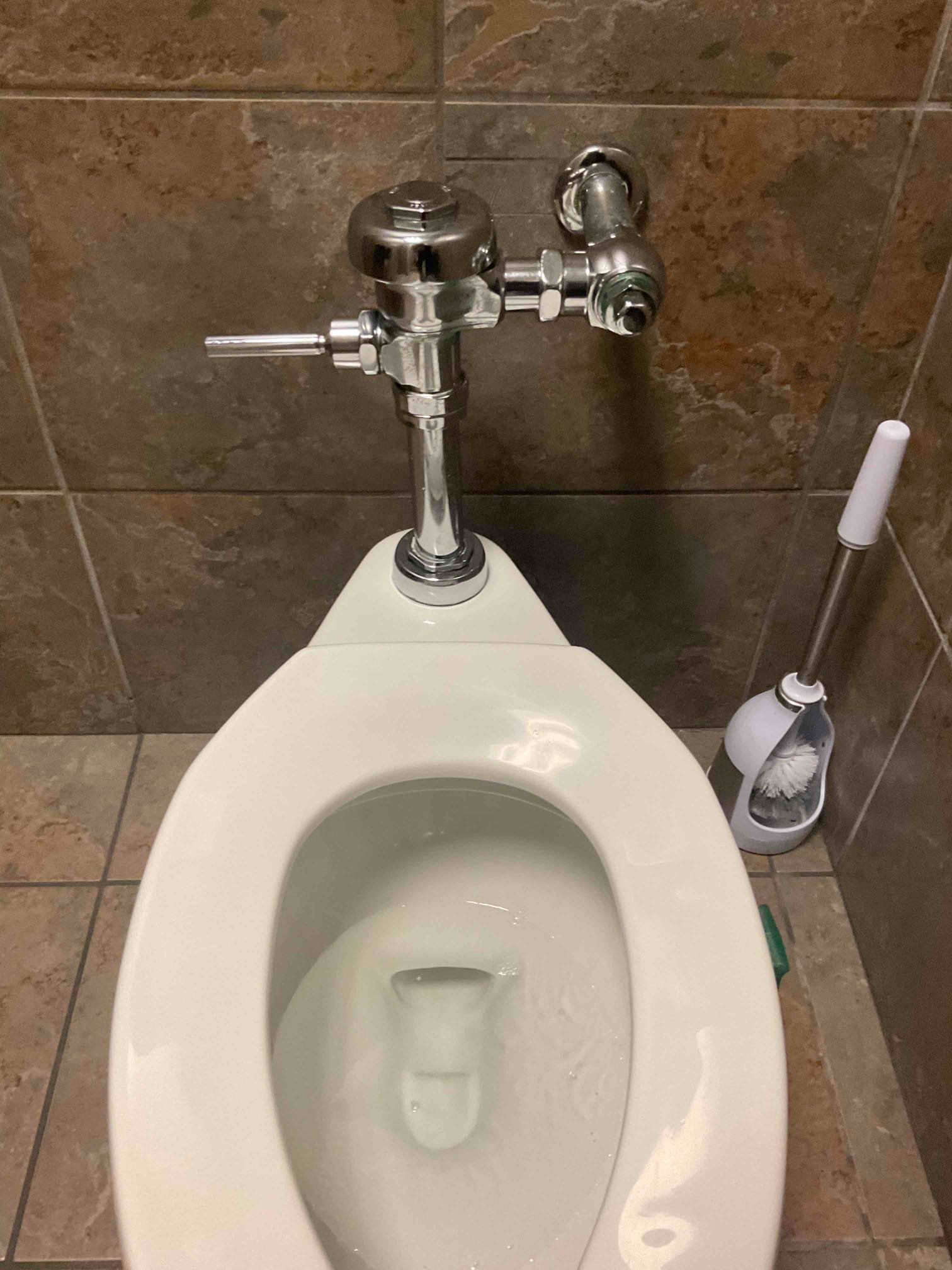 Urinal in a tiled restroom with a flush valve and toilet brush beside it