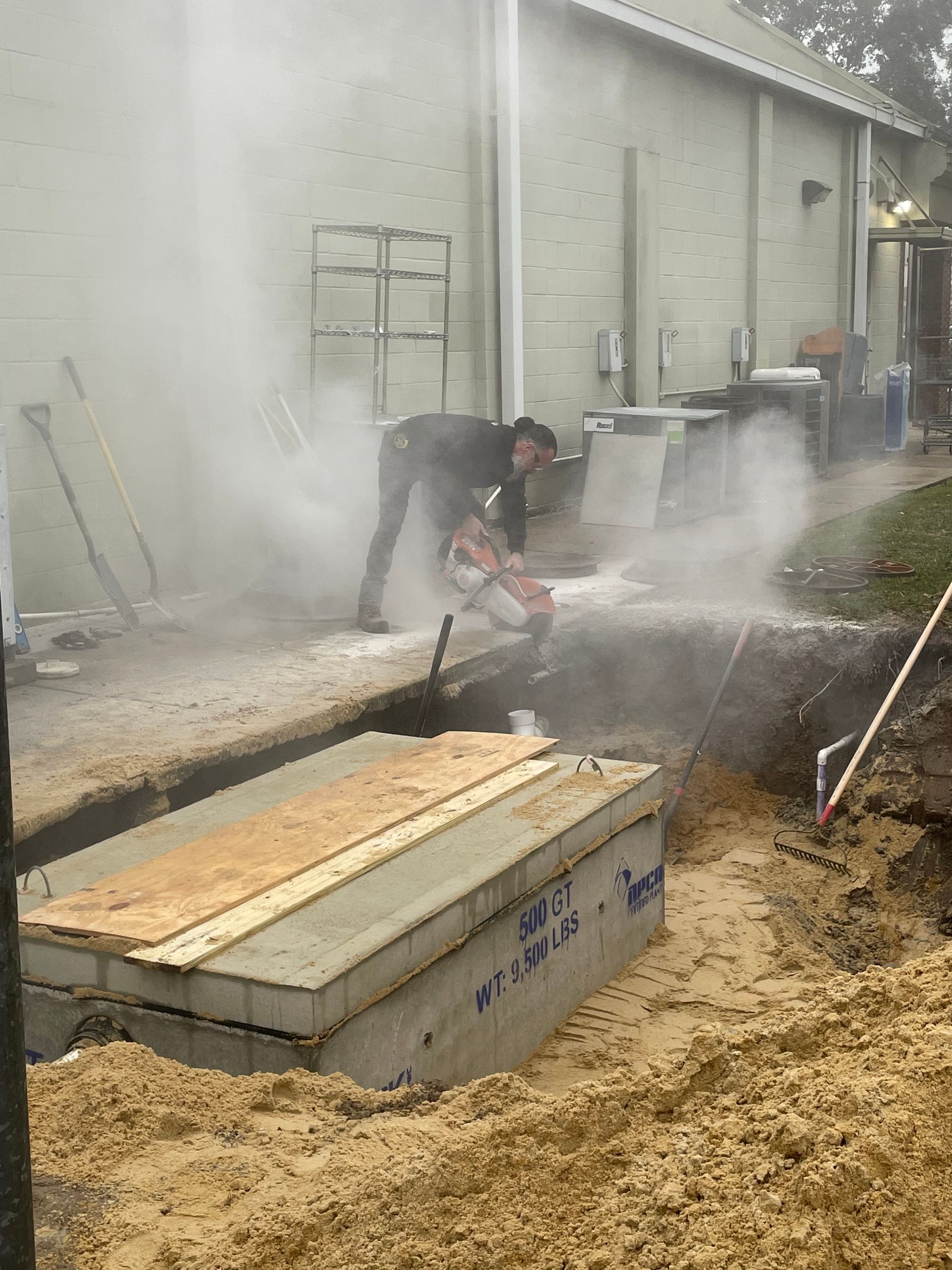 A worker uses a concrete saw to cut through a section of sidewalk next to an excavated trench.