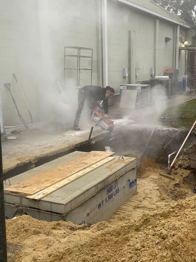A worker uses a concrete saw to cut through a section of sidewalk next to an excavated trench.