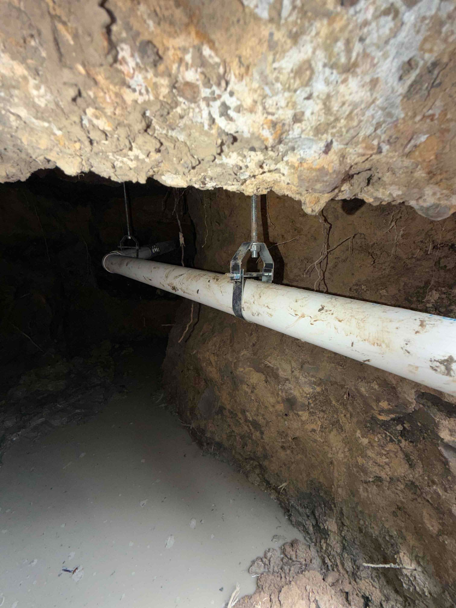 Underground tunnel with a white pipe running along a rough rock wall and muddy floor