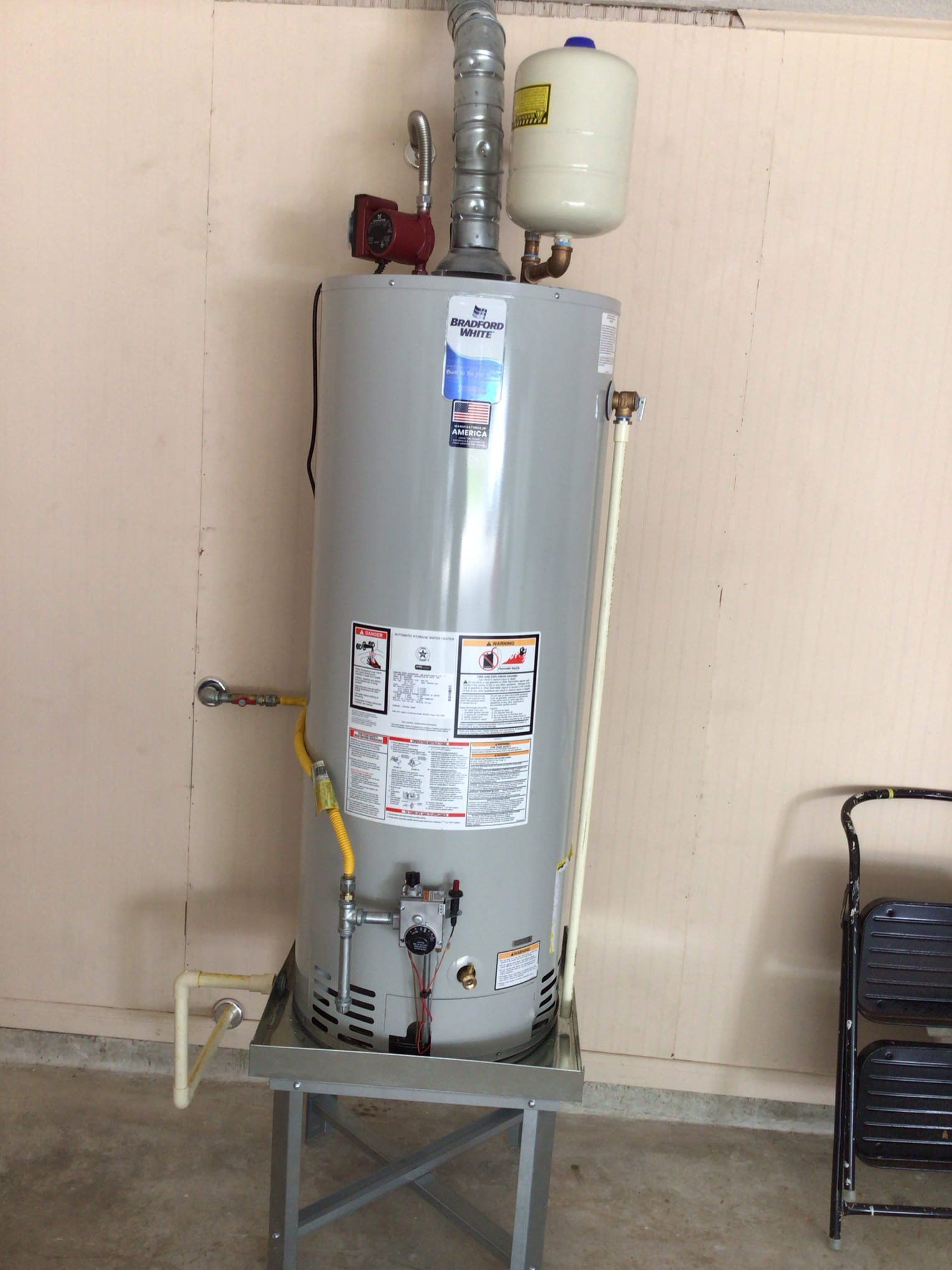 Gray water heater tank with pipes and a white expansion tank, standing on a metal stand indoors.