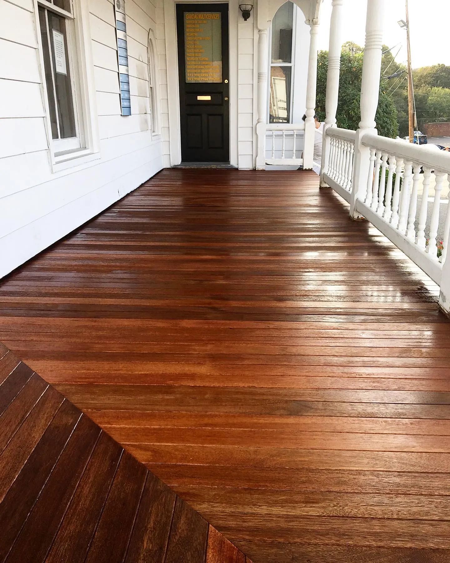 A wooden porch with newly stained floorboards. White house with black door and white railing.