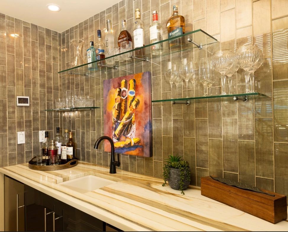 Bar area with glass shelves holding bottles, glassware, and a painting against a textured gold wall.