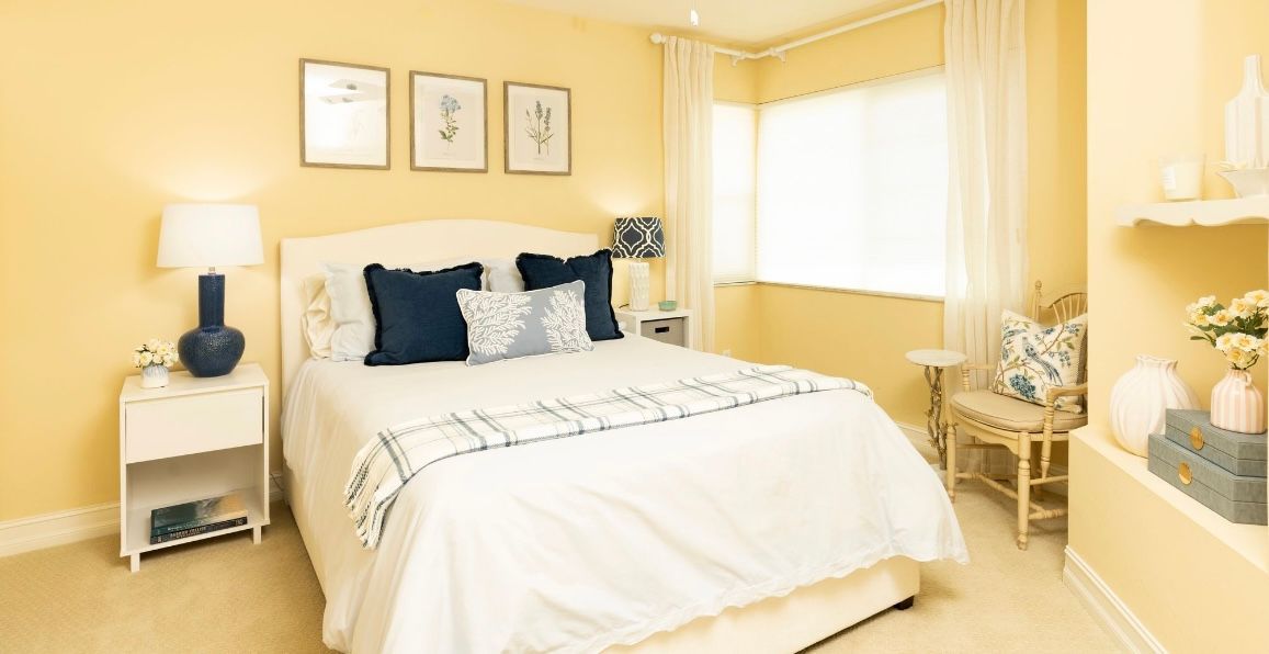 Yellow bedroom with white bed, blue pillows, and floral art.