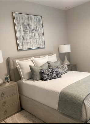 Bedroom with a beige bed, matching nightstands, and abstract artwork on the wall. The bed has white bedding and accent pillows in shades of gray.