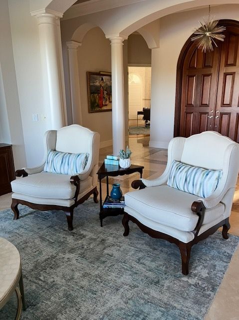 Two armchairs with blue pillows, small table between them, set on a blue rug, near a wooden door.