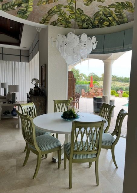 Dining room with a round white table, green chairs, and a decorative chandelier, near a window overlooking a patio.