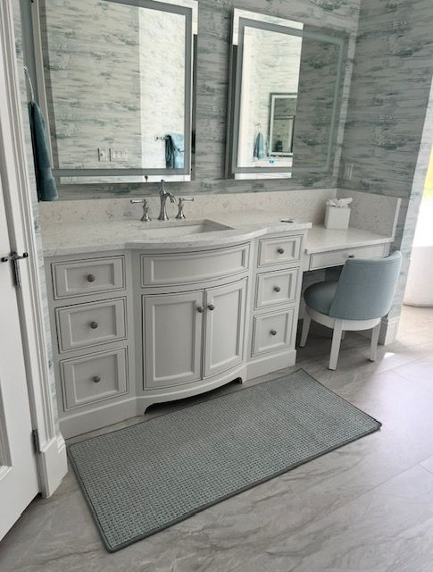 Bathroom with white vanity, gray rug, and blue chair. Mirrors reflect the marble-like wall.