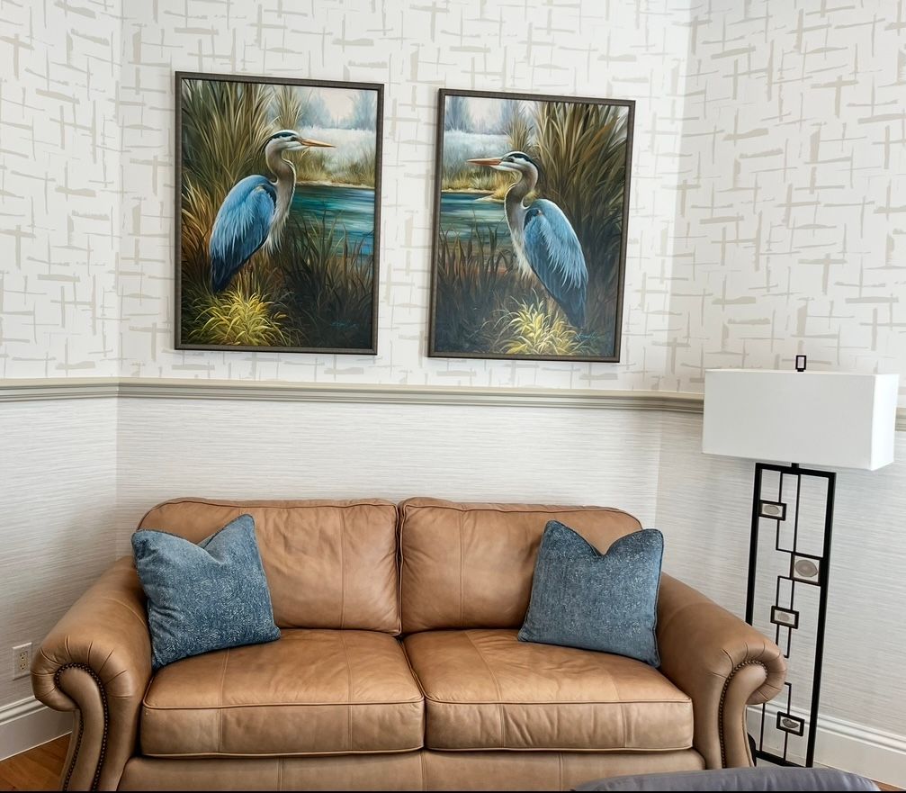 Two heron paintings above a brown leather sofa with blue pillows. Modern floor lamp beside sofa.
