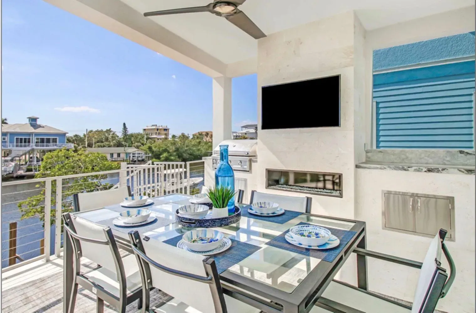 Outdoor dining area overlooking water with table set for meal, TV, grill, and blue accents.