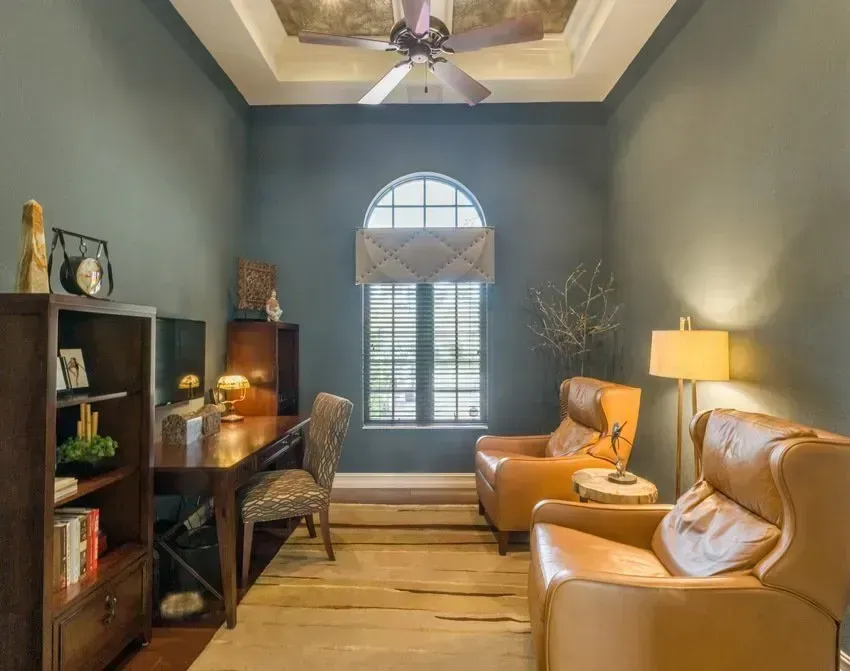 Home office with blue-green walls, desk, bookcase, two leather armchairs, and arched window with shuttered blinds.