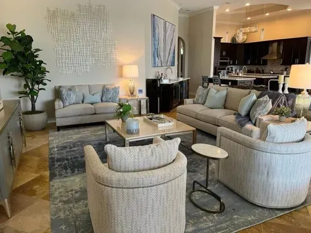 Living room with beige furniture, area rug, open to kitchen with dark cabinets.