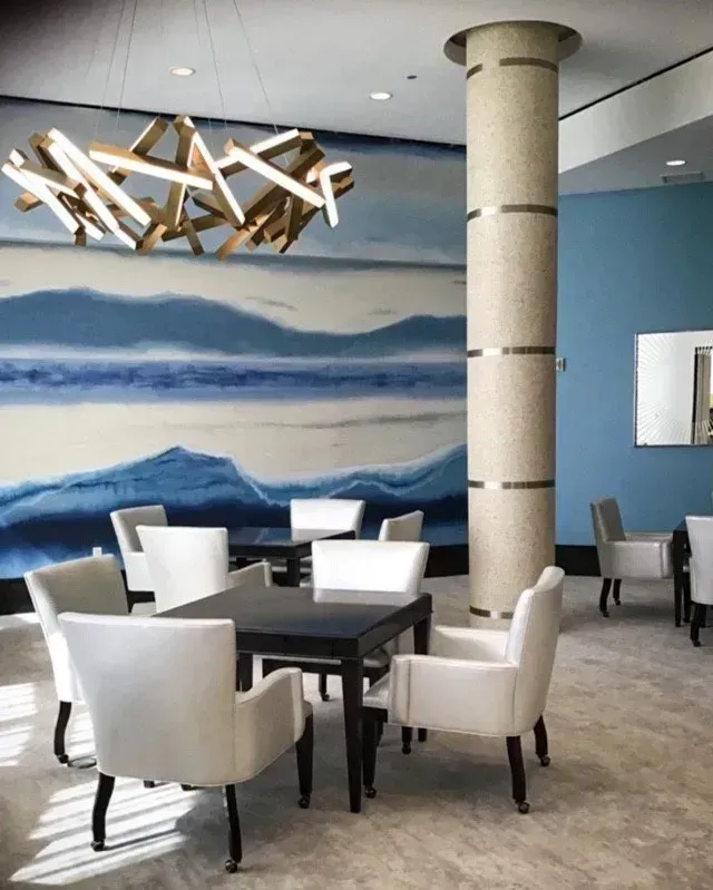 A lounge area with white chairs, black tables, and a light fixture against a blue mural, near a decorative pillar.