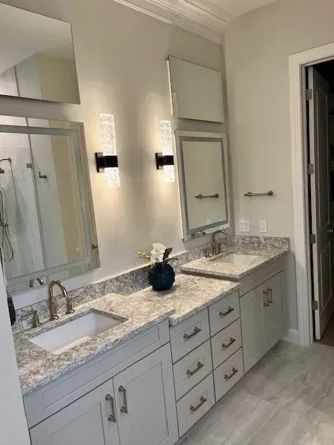 Bathroom with two sinks, gray cabinets, granite countertops, mirrors, and sconce lighting.