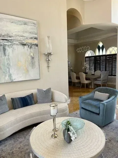 Elegant living room with curved white sofa, blue armchair, and artwork. Soft colors, arched doorway to dining area.