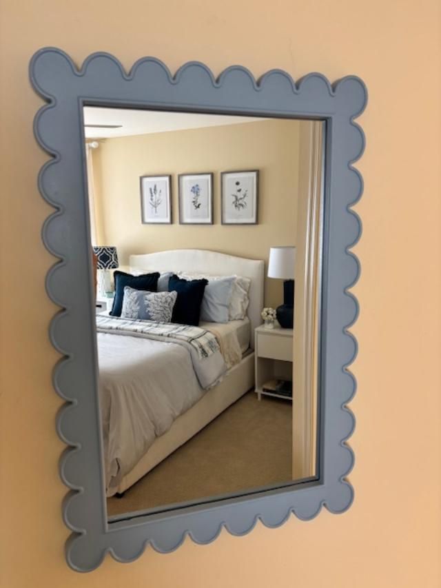 Scalloped blue-framed mirror reflecting a bedroom with a white bed, neutral walls, and artwork.
