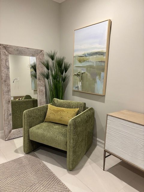 Green armchair with yellow pillow, mirror, artwork, side table, and floor plant in a room.