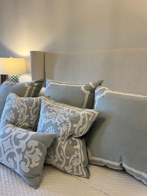Bed with light gray pillows, including decorative patterned and bordered cushions, against a neutral headboard.