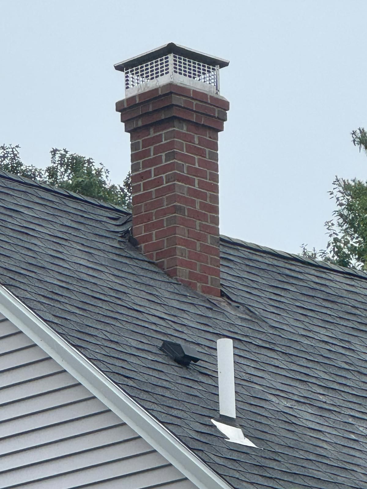 A brick chimney with a metal cap sits on a shingled roof, next to a white plumbing vent pipe.