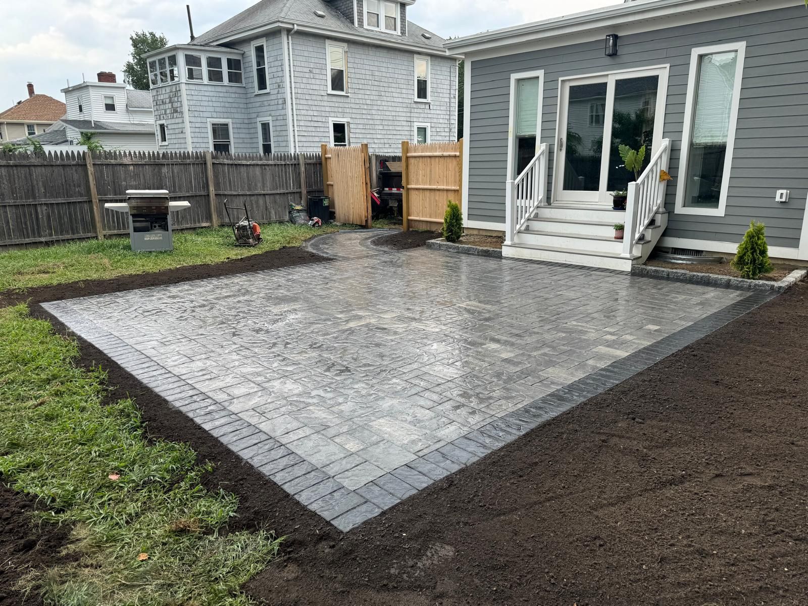 A newly installed gray paver patio with a dark border sits in a backyard next to a gray house with white steps.