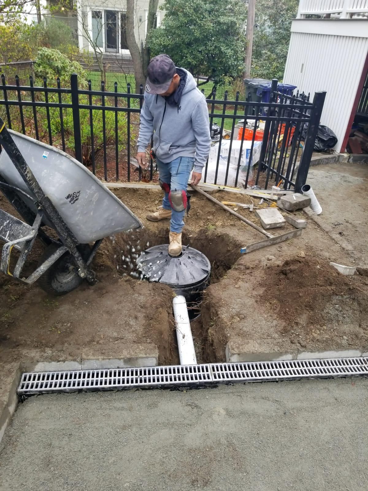 A person in work clothes stands over a drainage basin in a dirt yard, holding a wheelbarrow while installing pipe.