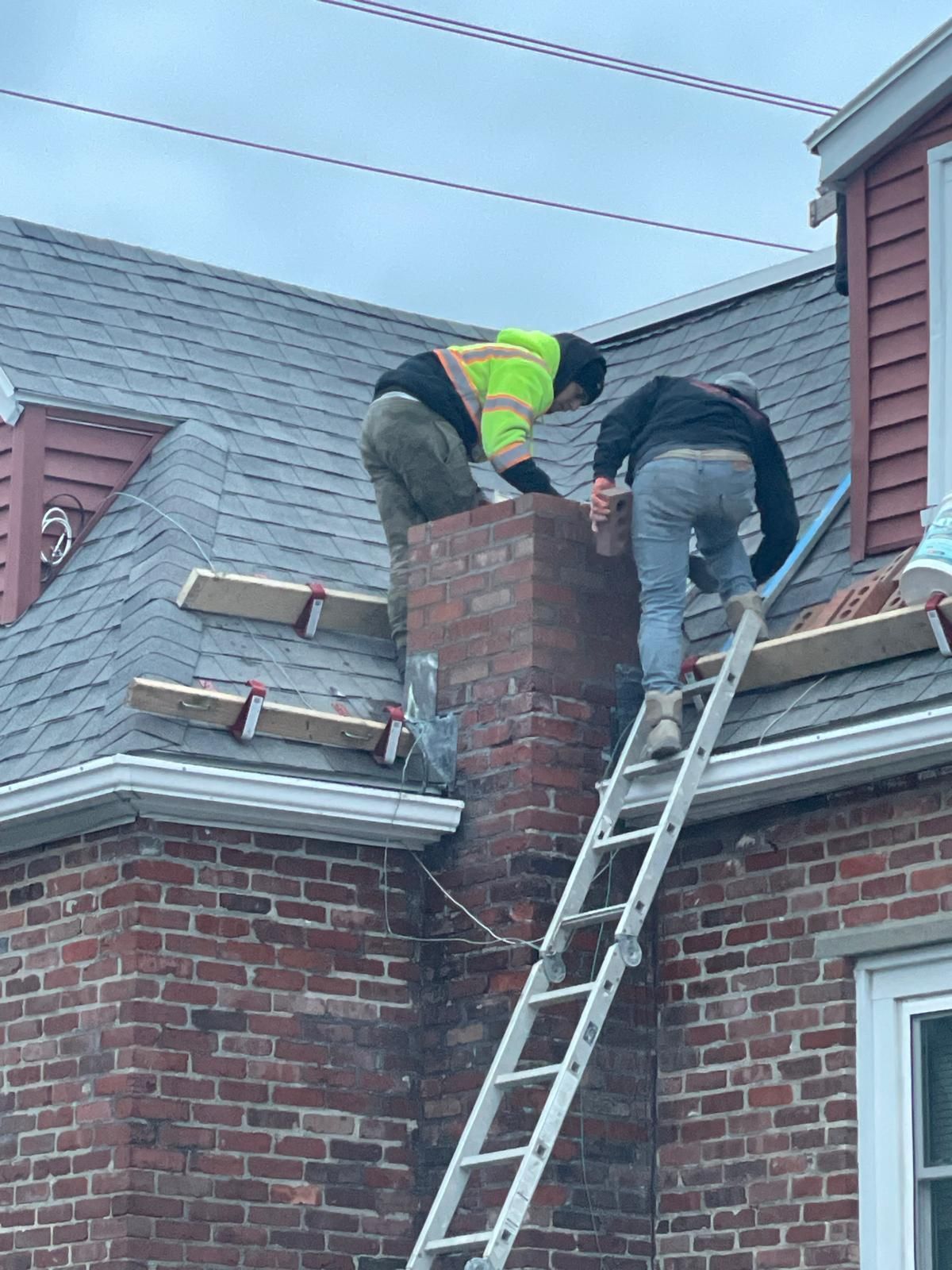 Two workers in work gear on a sloped roof repairing a brick chimney, with one worker climbing a ladder.