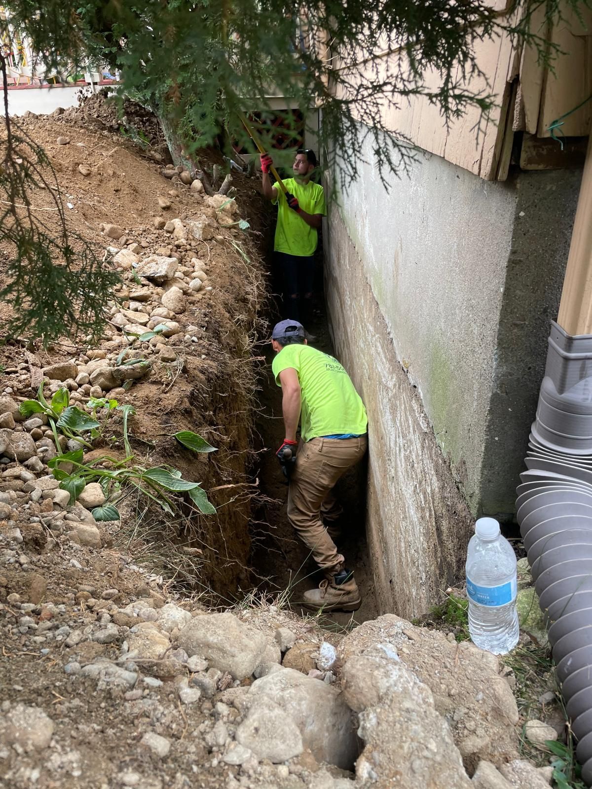 Two workers in neon yellow shirts dig a narrow trench along the foundation of a house.