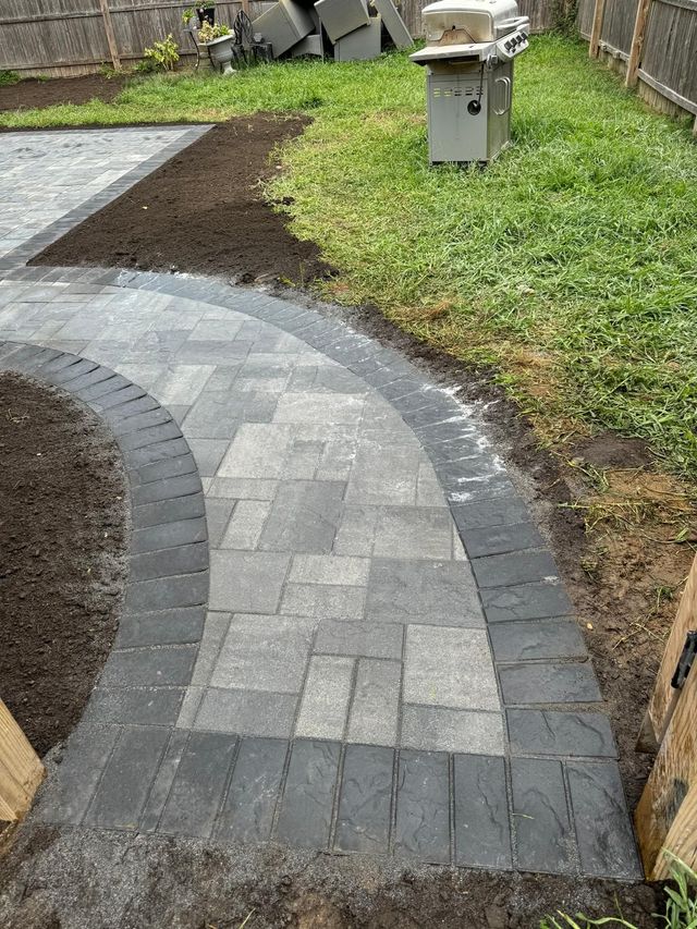 A curved gray paver walkway with a dark border cuts through a lawn next to a brown wooden fence and a metal grill.
