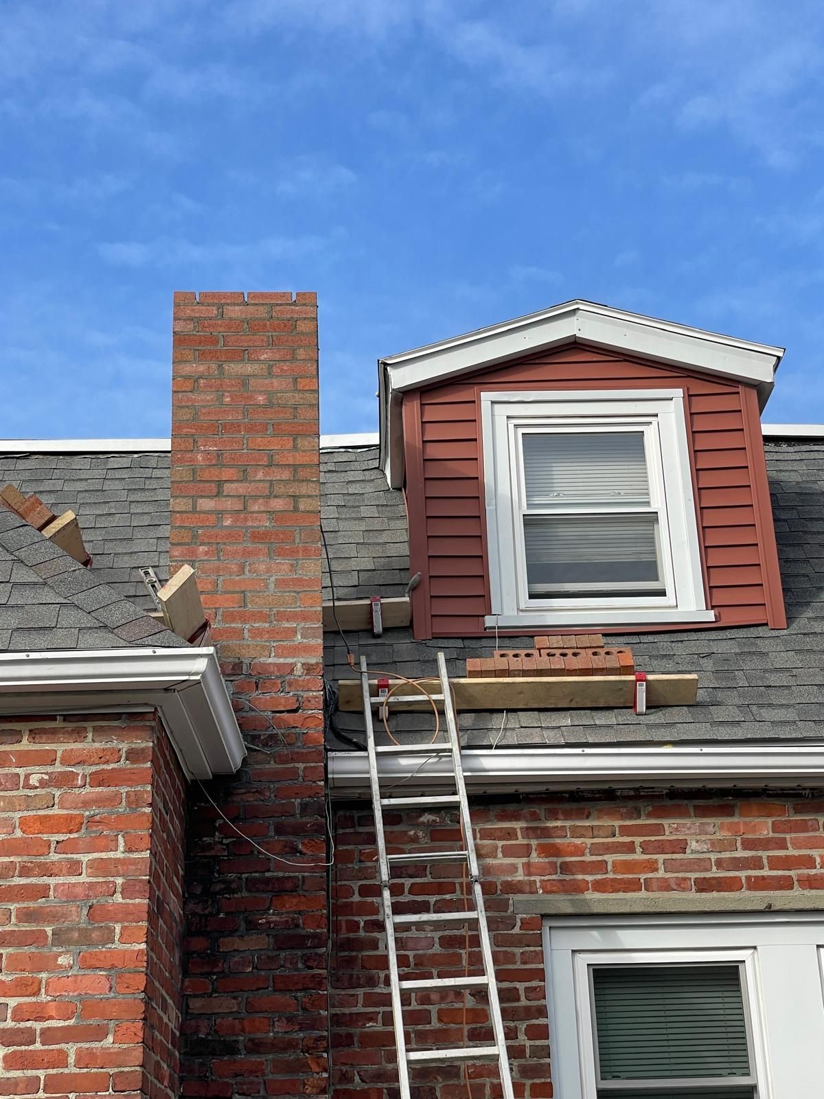 A ladder leans against a brick house roof next to a chimney and a small wooden scaffold platform under a dormer window.