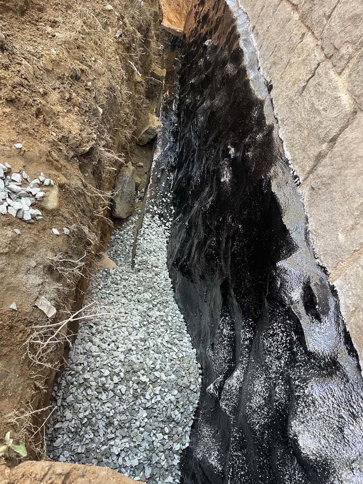 A deep trench along a foundation wall coated with black waterproofing, with a base layer of grey gravel.