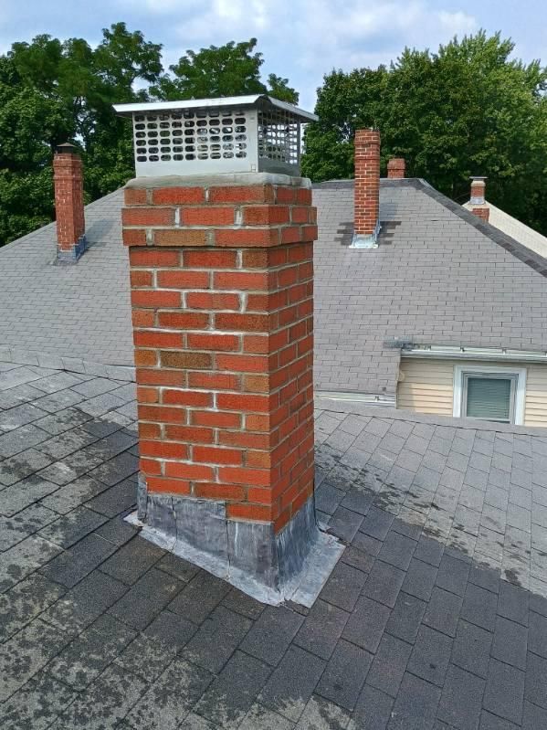 Red brick chimney with a metal cap and flashing on a gray shingled roof, surrounded by other chimneys and trees.
