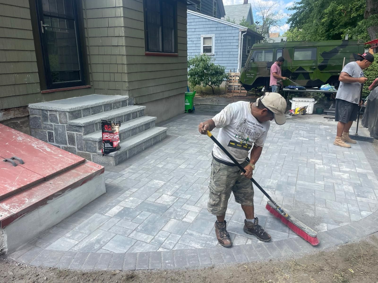 A worker in a t-shirt sweeps sand over newly laid gray stone pavers in front of a house entrance and brick stairs.