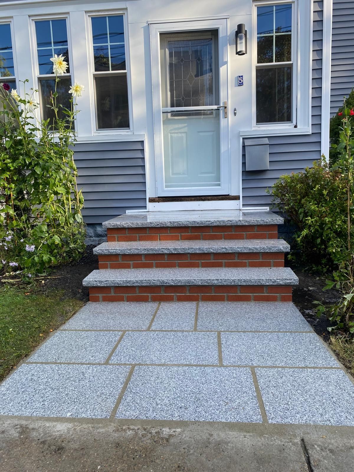 A front entry with a light grey, stone-tiled walkway and three matching stone-tiled steps with red brick risers.