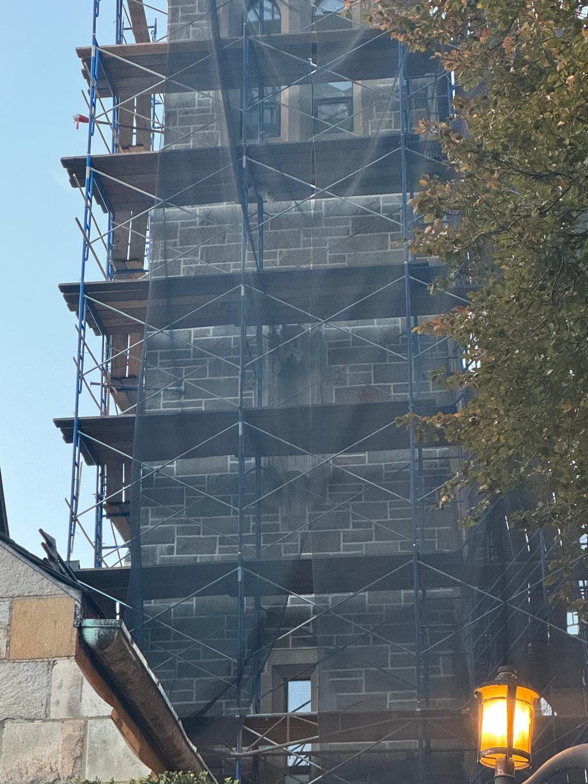 A tall stone tower under renovation, surrounded by blue scaffolding and black construction netting against a bright sky.