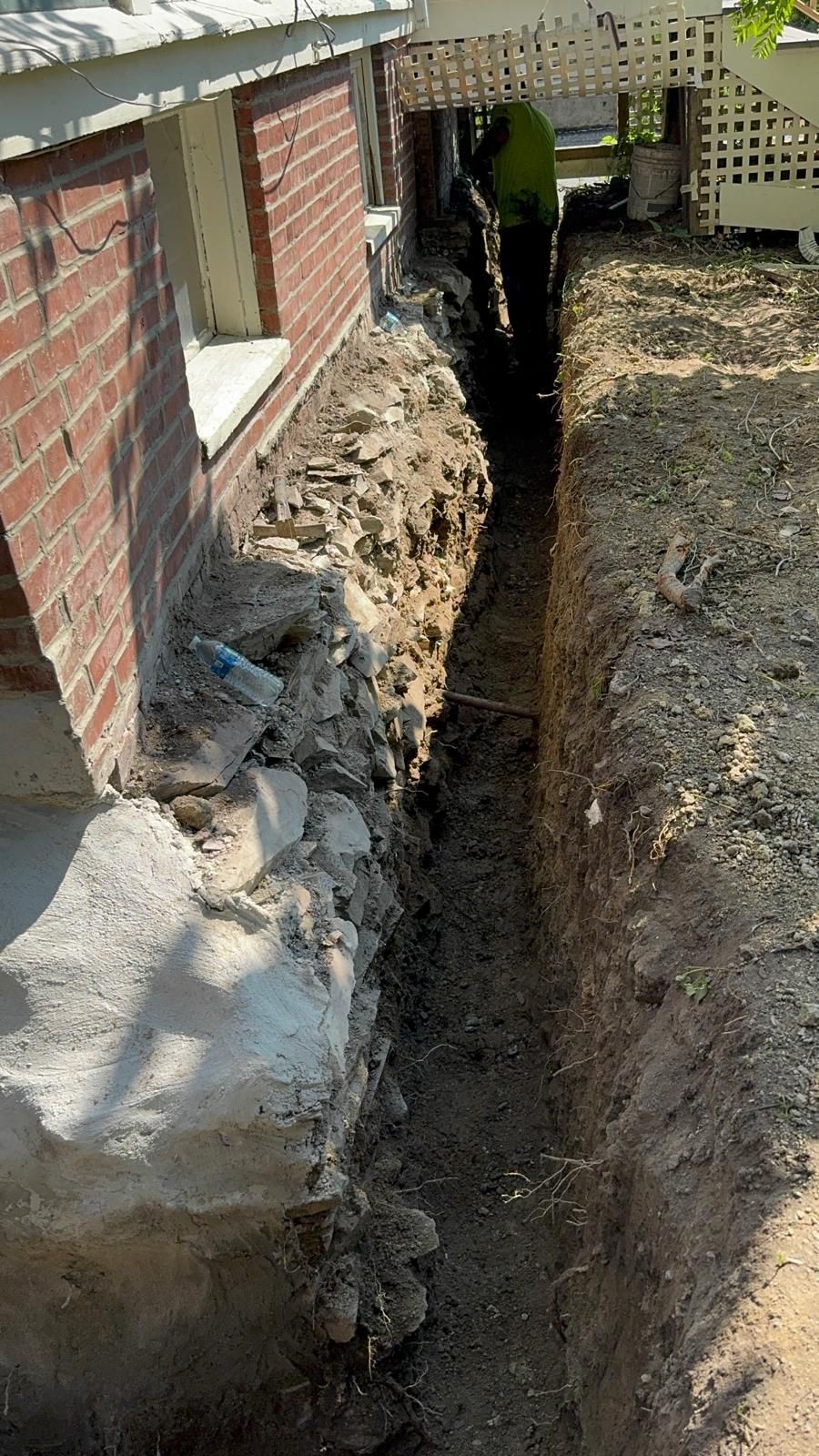 A trench dug alongside the brick foundation of a house, exposing the concrete base with a person visible at the far end.