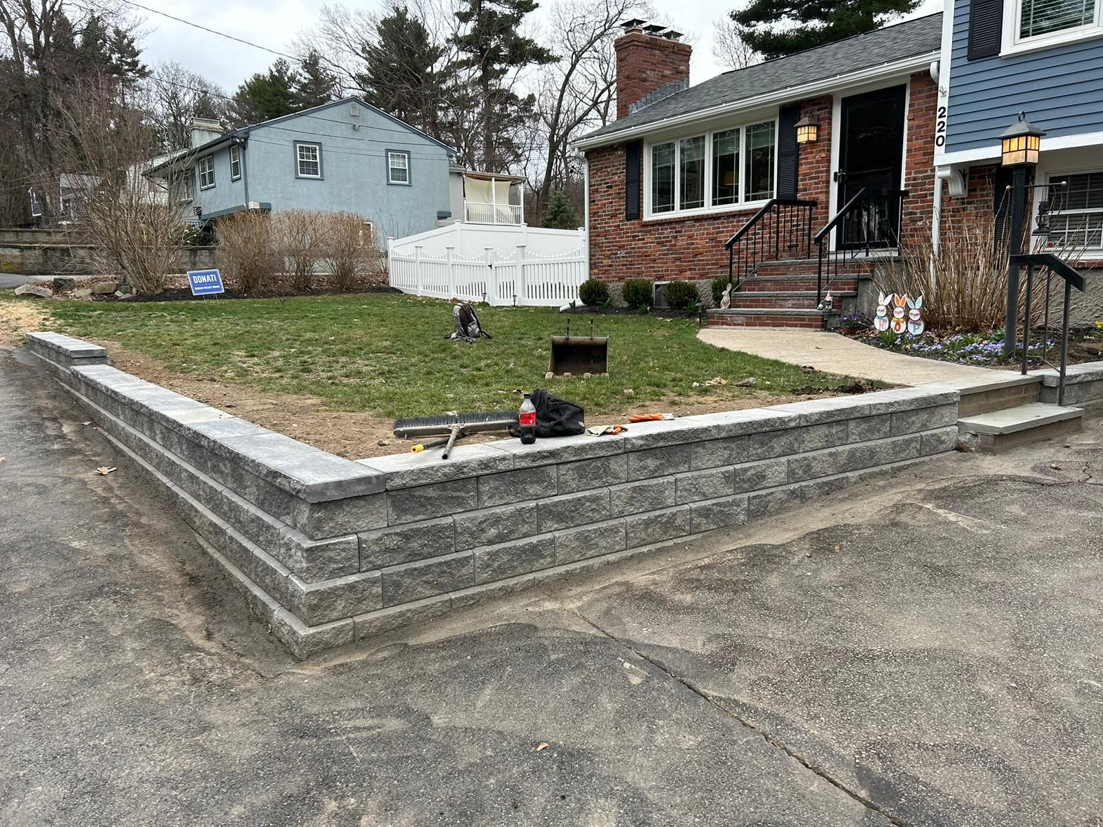 A newly constructed gray stone retaining wall separates an asphalt driveway from a grassy front yard of a brick house.