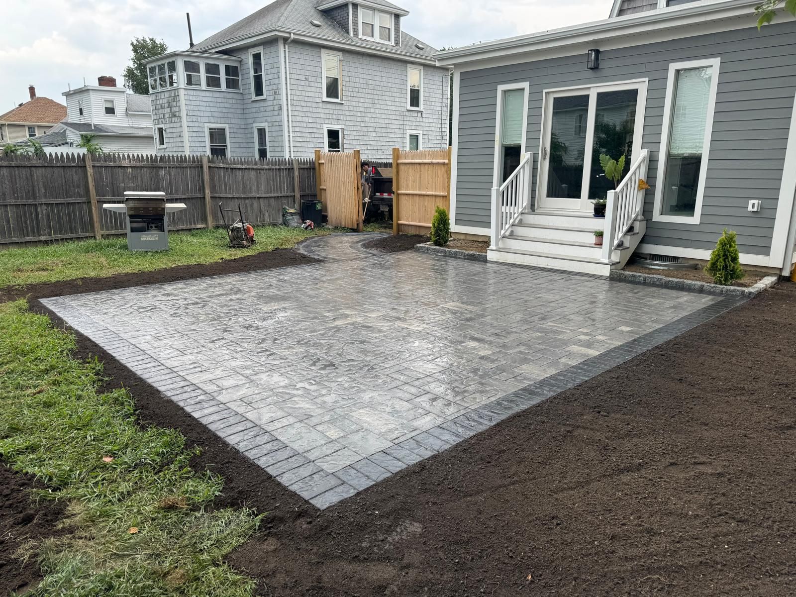 A newly installed rectangular stone paver patio next to a gray house with white stairs, surrounded by fresh dirt and grass.
