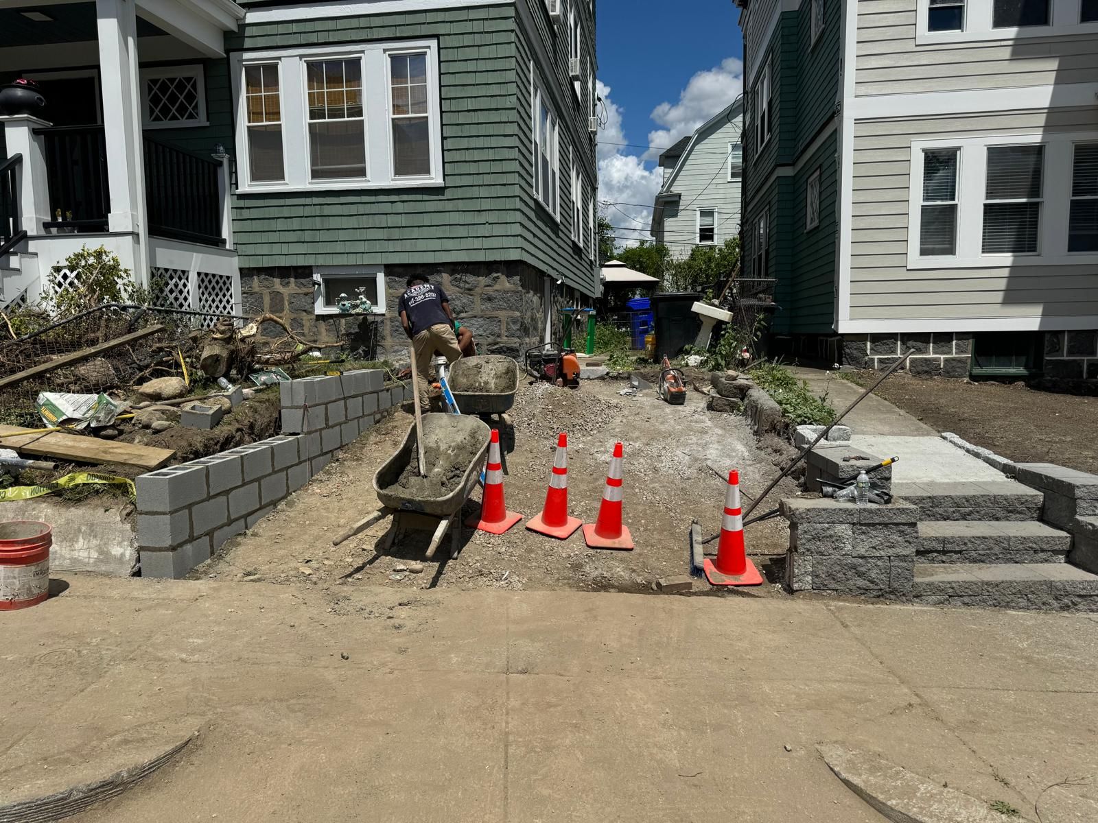 Construction workers installing a brick retaining wall and new steps between two residential houses on a sunny day.