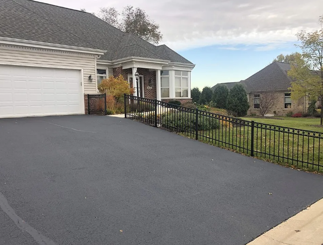 Black asphalt driveway slopes up to a house with a garage and entry.  A black iron fence lines the driveway's edge.