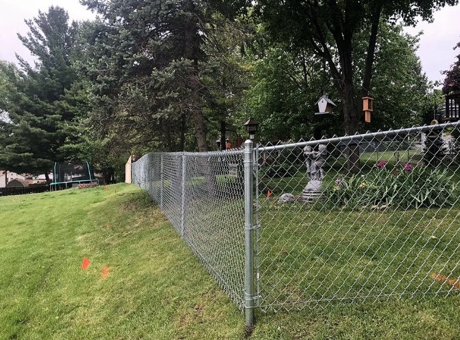 Chain-link fence on a grassy hill, separating the yard from trees and a house.
