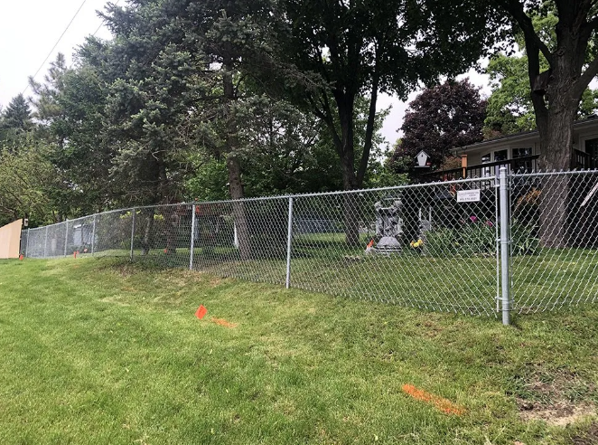 Chain link fence on a grassy hill; trees and a house in the background. Orange markers on the grass.