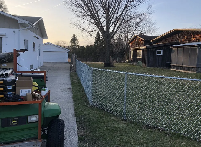 A chain-link fence separates a grassy yard from a concrete driveway, with a house, garage, and shed in the background.