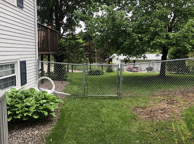 Chain-link fence encloses a green lawn near a house. A gate is open. Trees are in the background.