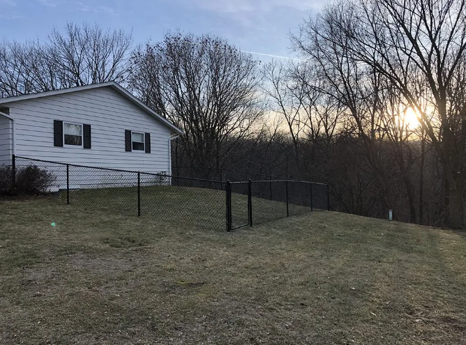 A black chain-link fence surrounds a grassy yard beside a white house; trees and sunset in background.