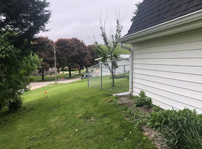 Side of white house next to green grass and a thin chain-link fence, trees in the distance.