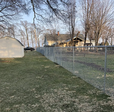 Chain-link fence in a yard, extending along a grassy area, with a house and shed in the background.