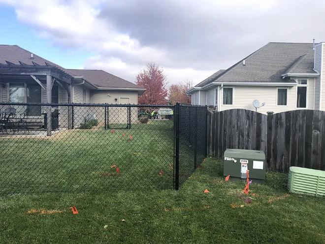 Backyard with chain link and wooden fences, utility boxes, green grass, and houses under a cloudy sky.