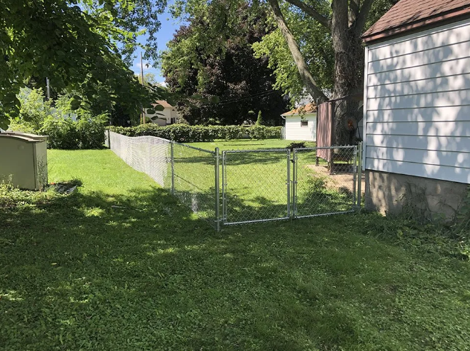 Backyard with chain-link fence and gate, partially enclosed, next to a white house with a shed on the left.