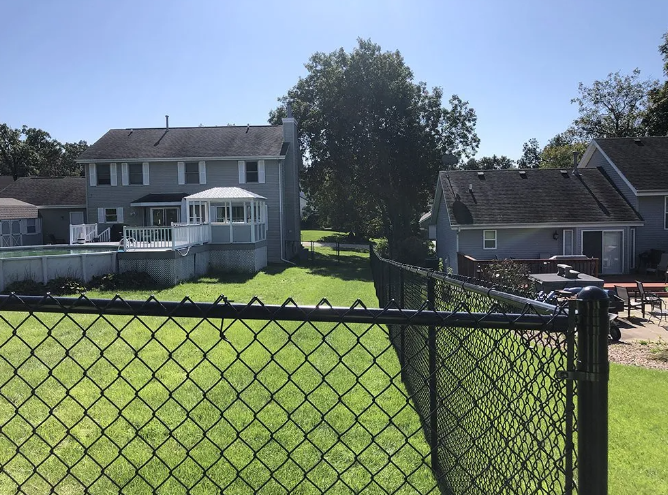 Black chain-link fence in front of green grass. Houses and trees are in the background under a blue sky.