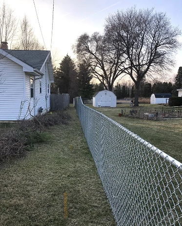 Chain-link fence in a backyard, beside a white house, on a grassy lawn.  Other structures and trees in the distance.